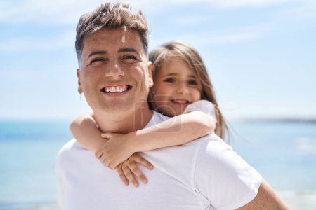 Téléchargez les photos : Père et fille souriant confiants s'embrassant tenant sur le dos au bord de la mer - en image libre de droit