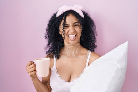 Téléchargez les photos : Femme hispanique avec des cheveux bouclés portant un pyjama boire une tasse de café coller la langue heureux avec une expression drôle. - en image libre de droit