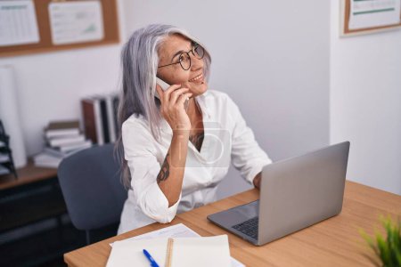 Foto de Trabajadora de negocios de mediana edad de cabello gris que usa computadora portátil que habla en el teléfono inteligente en la oficina - Imagen libre de derechos