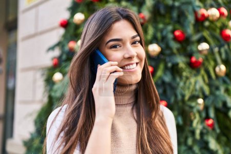 Téléchargez les photos : Jeune belle femme hispanique parlant sur smartphone debout près de l'arbre de Noël dans la rue - en image libre de droit