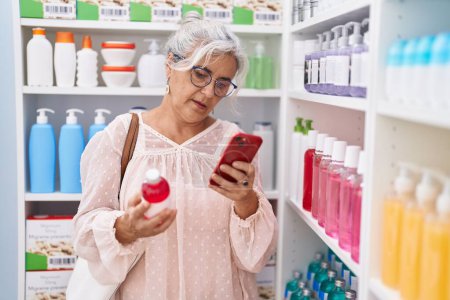 Photo for Middle age grey-haired woman customer using smartphone holding medicine bottle at pharmacy - Royalty Free Image