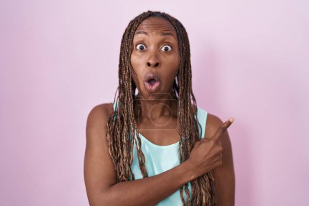 Foto de Mujer afroamericana de pie sobre fondo rosa sorprendida señalando con el dedo hacia un lado, boca abierta expresión asombrada. - Imagen libre de derechos
