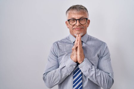 Foto de Hispanic business man with grey hair wearing glasses praying with hands together asking for forgiveness smiling confident. - Imagen libre de derechos