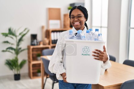 Téléchargez les photos : Femme afro-américaine travaillant au bureau tenant bouteille en plastique pour le recyclage clin d'oeil en regardant la caméra avec expression sexy, gai et heureux visage. - en image libre de droit
