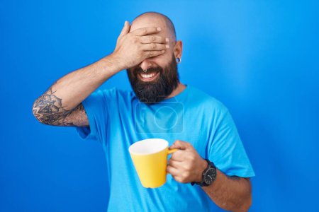 Foto de Young hispanic man with beard and tattoos drinking a cup of coffee smiling and laughing with hand on face covering eyes for surprise. blind concept. - Imagen libre de derechos