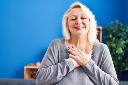 Téléchargez les photos : Femme blonde du moyen âge souriant confiant avec les mains sur le cœur à la maison - en image libre de droit