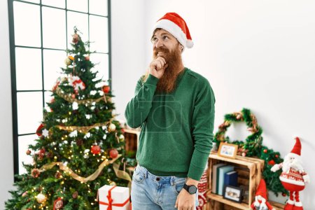 Téléchargez les photos : Homme rousse à longue barbe portant un chapeau de Noël par arbre de Noël avec la main sur le menton penser à la question, expression coûteuse. Visage souriant et réfléchi. concept de doute. - en image libre de droit