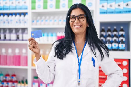 Téléchargez les photos : Femme hispanique mature travaillant à la pharmacie pharmacie tenant la carte de crédit regardant positif et heureux debout et souriant avec un sourire confiant montrant les dents - en image libre de droit