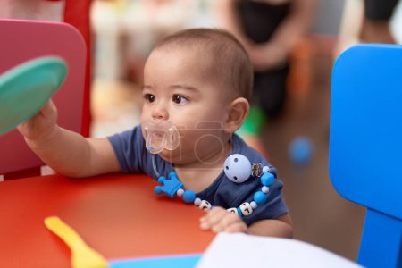 Photo for Adorable chinese toddler using pacifier holding dish toy at kindergarten - Royalty Free Image