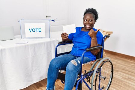 Téléchargez les photos : Jeune femme africaine assise en fauteuil roulant votant en mettant l'enveloppe dans l'urne souriant avec le visage heureux regardant et pointant vers le côté avec le pouce vers le haut. - en image libre de droit