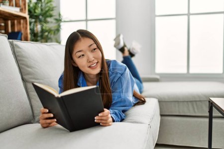 Photo for Young chinese girl reading book lying on the sofa at home. - Royalty Free Image