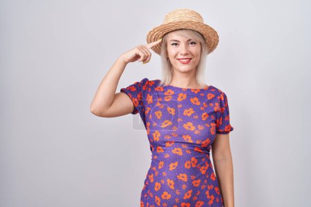 Foto de Mujer caucásica joven con vestido de flores y sombrero de verano sonriendo señalando a la cabeza con un dedo, gran idea o pensamiento, buena memoria - Imagen libre de derechos