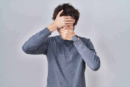 Foto de Young man standing over isolated background covering eyes and mouth with hands, surprised and shocked. hiding emotion - Imagen libre de derechos