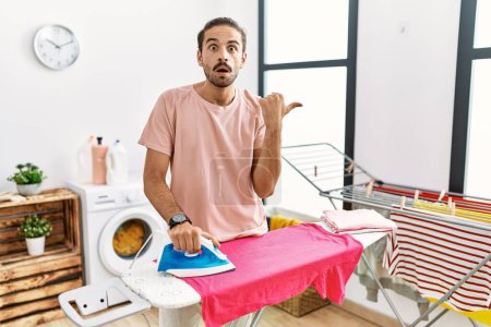 Téléchargez les photos : Jeune homme hispanique repassage vêtements à la maison surpris pointant avec le doigt de la main sur le côté, bouche ouverte expression étonnante. - en image libre de droit
