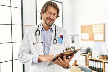 Foto de Middle age caucasian man wearing doctor uniform reading diagnosis al clinic - Imagen libre de derechos