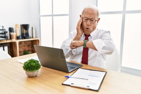 Téléchargez les photos : Homme âgé travaillant au bureau à l'aide d'un ordinateur portable en regardant l'heure de la montre inquiet, peur de se faire tard - en image libre de droit