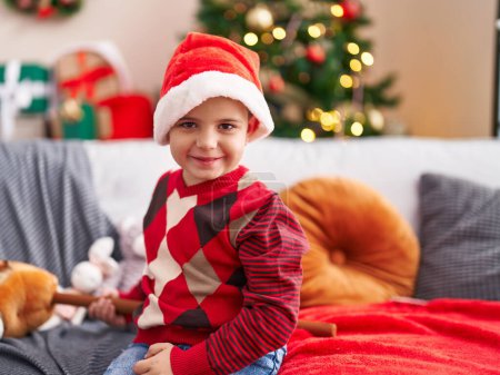 Foto de Adorable niño hispano sonriendo confiado sentado en el sofá por el árbol de Navidad en casa - Imagen libre de derechos