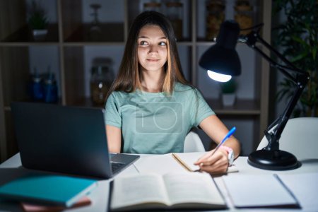 Foto de Adolescente chica haciendo la tarea en casa tarde en la noche sonriendo mirando a un lado y mirando hacia otro pensando. - Imagen libre de derechos
