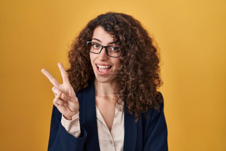Foto de Hispanic woman with curly hair standing over yellow background smiling with happy face winking at the camera doing victory sign. number two. - Imagen libre de derechos