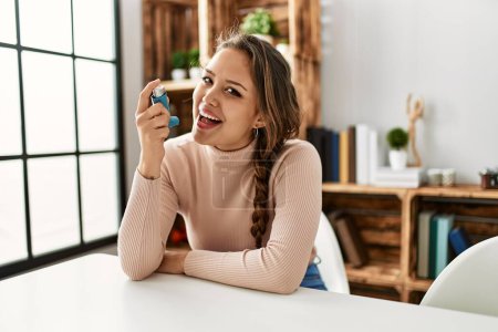 Photo for Young beautiful hispanic woman using inhaler sitting on table at home - Royalty Free Image