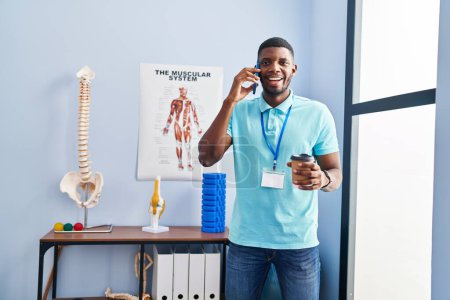 Téléchargez les photos : Young african american man physiotherapist talking on the smartphone and drinking coffee al rehab clinic - en image libre de droit