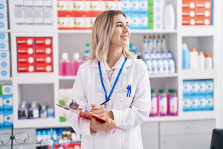 Téléchargez les photos : Young woman pharmacist smiling confident writing on document at pharmacy - en image libre de droit