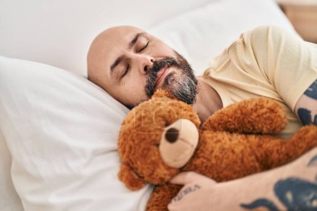 Photo for Young bald man hugging teddy bear lying on bed sleeping at bedroom - Royalty Free Image