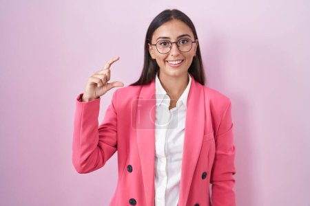 Téléchargez les photos : Young hispanic woman wearing business clothes and glasses smiling and confident gesturing with hand doing small size sign with fingers looking and the camera. measure concept. - en image libre de droit