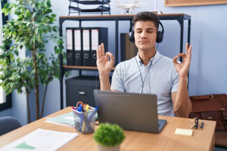 Téléchargez les photos : Jeune homme hispanique travaillant au bureau portant des écouteurs se détendre et sourire les yeux fermés en faisant geste de méditation avec les doigts. concept de yoga. - en image libre de droit
