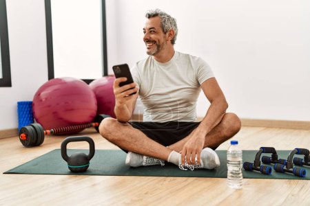 Foto de Hombre de pelo gris de mediana edad sentado en una esterilla de yoga usando un teléfono inteligente en el centro deportivo - Imagen libre de derechos