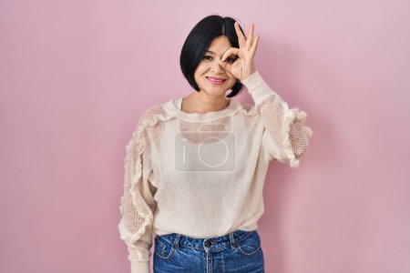 Téléchargez les photos : Young asian woman standing over pink background doing ok gesture with hand smiling, eye looking through fingers with happy face. - en image libre de droit