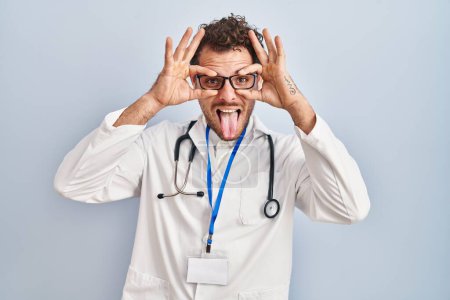 Photo for Young hispanic man wearing doctor uniform and stethoscope doing ok gesture like binoculars sticking tongue out, eyes looking through fingers. crazy expression. - Royalty Free Image