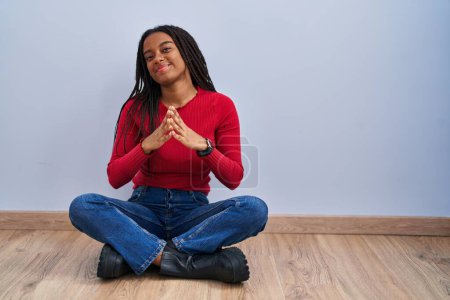Téléchargez les photos : Jeune afro-américain avec des tresses assises sur le sol à la maison les mains ensemble et les doigts croisés souriant détendu et joyeux. succès et optimisme - en image libre de droit