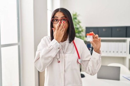 Photo for Young hispanic doctor woman holding sample cup covering mouth with hand, shocked and afraid for mistake. surprised expression - Royalty Free Image