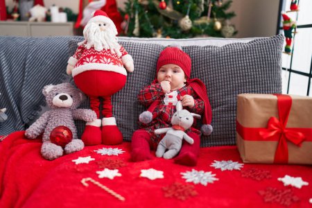 Foto de Adorable bebé caucásico chupando el dedo sentado en el sofá por el árbol de Navidad en casa - Imagen libre de derechos