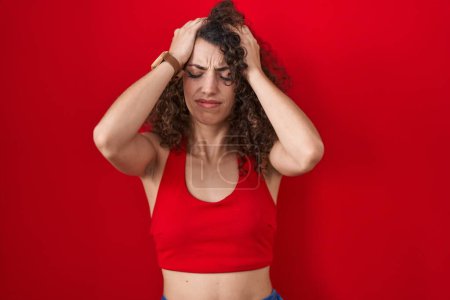 Foto de Mujer hispana con el pelo rizado de pie sobre el fondo rojo que sufre de dolor de cabeza desesperada y estresada porque el dolor y la migraña. manos en la cabeza. - Imagen libre de derechos
