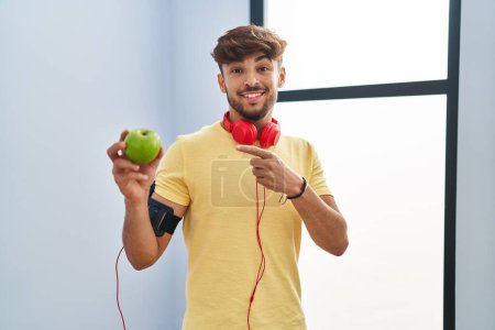 Photo for Arab man with beard wearing sportswear eating green apple smiling happy pointing with hand and finger - Royalty Free Image
