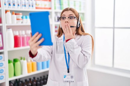 Téléchargez les photos : Redhead woman working at pharmacy drugstore doing video call with tablet covering mouth with hand, shocked and afraid for mistake. surprised expression - en image libre de droit