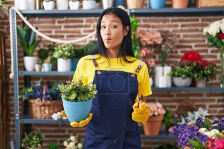 Photo for Hispanic woman working at florist shop holding plant making fish face with mouth and squinting eyes, crazy and comical. - Royalty Free Image