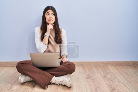 Photo for Young brunette woman working using computer laptop sitting on the floor thinking worried about a question, concerned and nervous with hand on chin - Royalty Free Image