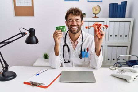 Foto de Young hispanic doctor man holding female genital organ and birth control pills smiling and laughing hard out loud because funny crazy joke. - Imagen libre de derechos