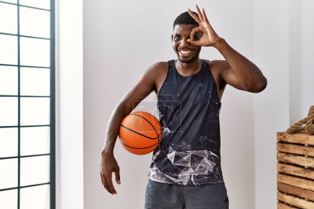 Foto de Joven afroamericano hombre sosteniendo pelota de baloncesto en casa sonriendo feliz haciendo signo bien con la mano en el ojo mirando a través de los dedos - Imagen libre de derechos