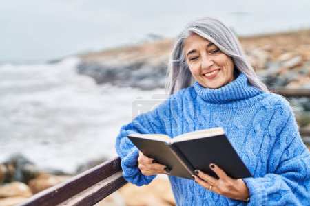Téléchargez les photos : Femme aux cheveux gris d'âge moyen souriant livre de lecture confiant au bord de la mer - en image libre de droit