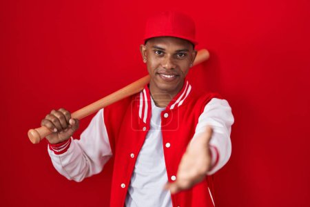 Téléchargez les photos : Jeune homme hispanique jouant au baseball tenant chauve-souris souriant offrant main de paume donnant assistance et acceptation. - en image libre de droit
