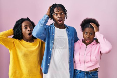 Foto de Grupo de tres jóvenes negros de pie juntos sobre fondo rosa locos y asustados con las manos en la cabeza, asustados y sorprendidos de shock con la boca abierta - Imagen libre de derechos