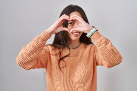 Téléchargez les photos : Jeune femme brune debout sur fond blanc faisant forme de coeur avec la main et les doigts souriant en regardant à travers signe - en image libre de droit