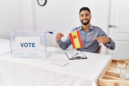 Foto de Joven hombre guapo con barba en las elecciones de campaña política con bandera de España sonriendo feliz señalando con la mano y el dedo - Imagen libre de derechos