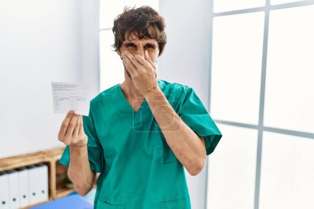 Photo for Young hispanic doctor man holding covid record card smelling something stinky and disgusting, intolerable smell, holding breath with fingers on nose. bad smell - Royalty Free Image