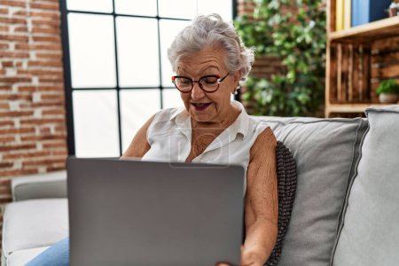 Téléchargez les photos : Femme âgée aux cheveux gris utilisant un ordinateur portable assis sur le canapé à la maison - en image libre de droit