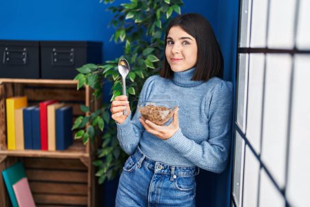 Photo for Young hispanic woman eating healthy whole grain cereals with spoon smiling looking to the side and staring away thinking. - Royalty Free Image